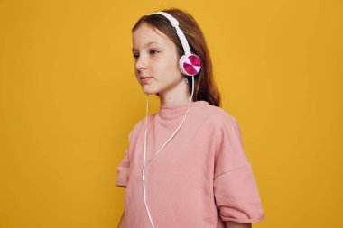 Pembe kazaklı genç kız sarı arka planda beyaz ve pembe kulaklıklı müzik dinliyor. Sakin bir ruh hali ve gündelik stili gösteren yan profil portresi.