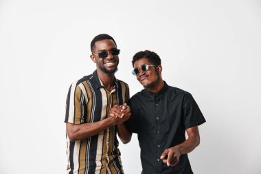 Siyah tenli, güneş gözlüklü ve günlük giysili Afro-Amerikalı erkekler el sıkışıyor ve dostluk ve birlik için beyaz arka planda birlikte gülümsüyorlar..