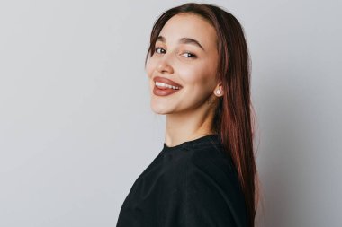 Siyah bluzlu, uzun düz saçlı, gülümseyen kadın beyaz duvarın önünde duruyor. Küpeli ve doğal görünüşlü neşeli bir kadının yakın plan portresi pozitif duyguyu gösteriyor..