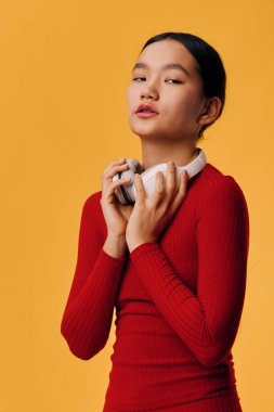 Kulaklık takan, sarı arka planda dikilen, gündelik kırmızı bluz giyen bir kadın model. Yüzünde sakin bir ifade, elleri boynuna yakın ses ekipmanı tutuyor..