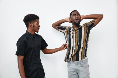 Çizgili ve siyah tişörtlü siyah tenli Afro-Amerikalı erkekler duygusal etkileşim içindeler. Bir adam kafasını şokta tutarken, diğeri beyaz zemin üzerine ciddi bir tartışmada uzanıyor..