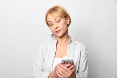 Sarışın, orta yaşlı, beyaz gömlekli, elinde akıllı telefon olan ve aşağıya bakan bir kadın. Beyaz arka plan stüdyo resmi. Dijital iletişim, günlük rutin ve teknoloji kullanımı kavramı.