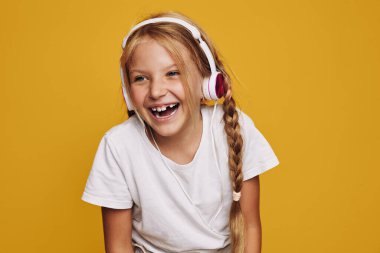 Saçları örgülü gülümseyen genç kız kulaklıkla müzik dinliyor. Parlak sarı arka plan ve beyaz tişört neşeli ve canlı bir hava yaratıyor. Çocuklar ve eğlence için kavram.