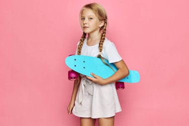 Sarı saçlı, örgülü genç kız elinde turkuaz bir kaykay tutuyor. Beyaz bir tişört ve bej bir etek giyiyor. Pembe stüdyo geçmişine karşı duruyor..
