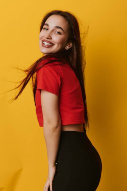 Kırmızı tişört ve siyah pantolon giyen, sarı arka planda saçları akan gülümseyen bir kadın. Olumlu duygu ve tarz gösteren neşeli genç bir kadının portresi..