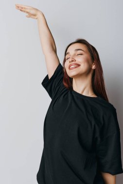 Kolunu kaldırmış gülümseyen ve siyah tişörtlü kapalı gözlü bir kadın tarafsız arkaplanda duruyor. Neşenin, güvenin ve doğal güzellik ve mutluluğun ifadesi..