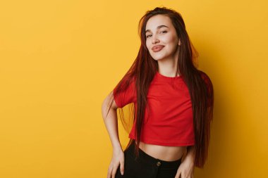 Uzun saçlı, kırmızı bluzlu, siyah pantolonlu, sarı arka planda duran kendine güvenen bir kadın. Sıradan moda, gülümseyen ifade ve modern portre için rahat poz..