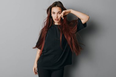 Gri duvarın önünde poz veren siyah tişörtlü, uzun saçlı genç bir kadın portresi. Kendine güveni olan bir kadın, eli başının yanında ve modaya uygun bir şekilde içeri bakar..