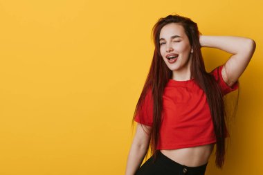 Kırmızı tişörtlü, uzun saçlı, gözleri kapalı poz veren ve elleri sarı arka planda olan bir kadın. Genç, rahat moda, doğal ifade ve rahat duruş..