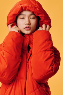 Kapüşonlu parlak turuncu yastıklı ceket giyen genç bir kadın, elleri tasmalı, gözleri kapalı. Stüdyo portresi, kışlık kıyafet, kalın dış giysi, rahat kıyafet, mevsimlik moda ve günlük tarz..