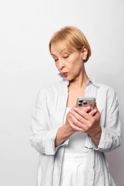 Beyaz gömlek ve pantolon giyen sarışın kadın akıllı telefonu iki eliyle tutuyor ve odaklanmış bir ifadeyle yana bakıyor. Teknoloji kullanımı, iletişim ve merak kavramı.