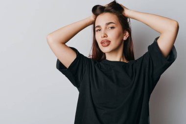 Uzun kahverengi saçlı, sıradan siyah tişörtlü, elleri başının üstünde poz veren genç bir kadının portresi. Tarafsız arkaplanlı stüdyo çekimi kendine güvenen ve sakin bir tavır sergiliyor.