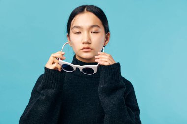 Siyah balıkçı yaka kazak giyen, beyaz gözlüklü, sakin, genç bir kadın mavi arka planda kapalı gözlerle poz veriyor. Doğal görünüşlü ve siyah saçlı bir kadın modelin stüdyo fotoğrafı..