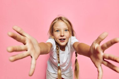 Beyaz tişörtlü, sarı örgülü heyecanlı bir çocuk pembe bir arka plana doğru ellerini kameraya doğru uzatıyor. Parlak stüdyo ışıkları canlı ve neşeli bir atmosfer yaratır..