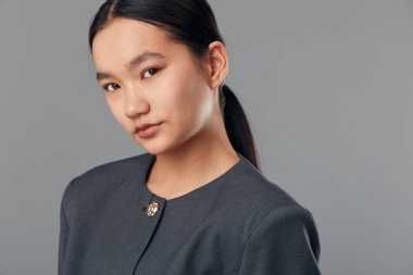 Sade at kuyruklu, koyu gri takım elbiseli, kendine güvenen Asyalı genç bir kadının portresi. Profesyonel vesikalık fotoğrafında gri zemine karşı duruyor..