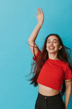 Kırmızı bluzlu ve siyah pantolonlu gülümseyen genç bir kadın mavi arka planda dalgalı saçlarıyla el kaldırıyor. Mutlu bir kadın portresi, stüdyoda doğal mutluluk ve gündelik stil..