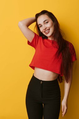 Kırmızı tişörtlü, uzun saçlı, siyah pantolonlu, sarı arka planda el üstünde poz veren, sıradan moda ve mutlu bir ifade sergileyen gülümseyen kadın..