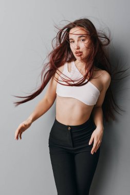 Uzun kahverengi saçlı, beyaz kolsuz bluzlu, siyah, geniş bel pantolonlu, gri arka planda, dinamik saç hareketli ve rahat görünümlü bir model..