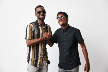 Siyah tenli, güneş gözlüklü Afro-Amerikalı erkekler el sıkışıyor ve beyaz arka planda günlük kıyafetlerle gülümsüyorlar. Arkadaşlık ve pozitif etkileşim için..