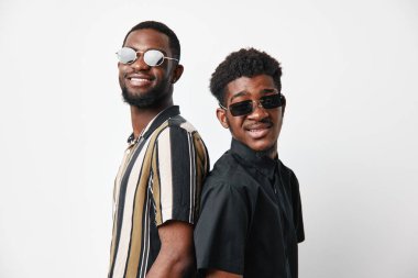 Siyah tenli, güneş gözlüklü ve günlük elbiseli iki Afrikalı adamın portresi. Stüdyodaki beyaz, pürüzsüz arka planda sırıtarak poz veriyor..