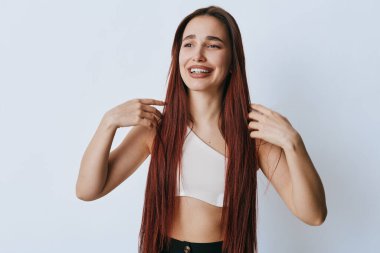 Uzun örgülü, gülümseyen genç bir kadın beyaz arka planda saçlarını düzeltiyor. Güzellik portresi, gündelik moda, mutluluk ve doğal cilt bakımı ve stüdyoda rahat duruş..