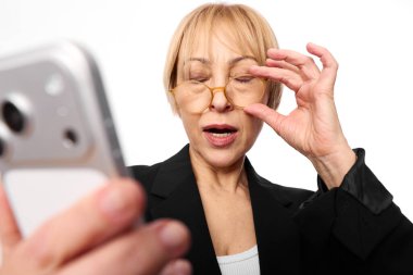 Gözlüklü ve siyah ceketli olgun bir kadın akıllı telefonu kapalı tutarken beyaz arka planda izole bir şekilde açık ağızla tepki veriyor. Sürpriz, teknoloji ve duygular için kavram.