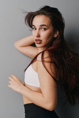 Portrait of young female model with flowing hair wearing casual white top and dark pants. Elegant pose, natural look, minimalistic background, and soft light for fashion or beauty concept.