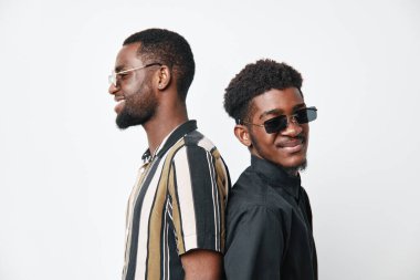 Siyah tenli siyah tenli ve güneş gözlüklü Afro-Amerikalı erkekler sırt sırta verip, stüdyoda güvenle gülümsüyorlar. Yaşam tarzı konsepti için beyaz arka planı var..