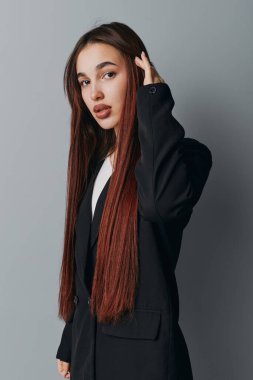 Uzun kahverengi saçlı, siyah ceketli, gri arka planda duran kadın model. Kameraya bakan düz saçlı ve ince makyajlı genç bir kadının yakın plan portresi..
