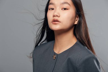 Gri ceketli genç Asyalı kadın, doğal makyaj ve uzun saçla güven içinde poz verdi. Moda, güzellik ve profesyonel portre konsepti için gri arka planda izole edildi..