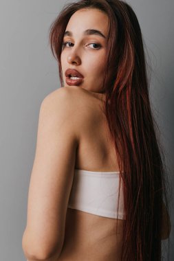 Uzun kahverengi saçlı, çıplak omuzlu, askısız bluz giyen genç bir kadının portresi. Doğal görünüş, tarafsız arka planda poz veren yumuşak ışıklandırma ve rahat ruh hali..