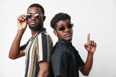 Sırt sırta duran güneş gözlüklü, koyu tenli iki siyah adamın portresi. Bir puan, günlük kıyafetler, çizgili ve siyah gömlekler, özgüvenli ifadeler ve stil ile stüdyo çekimi.