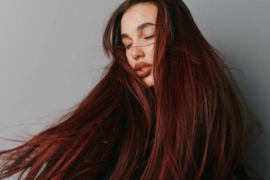 Canlı kızıl saçlı, gevşek yüzlü, düz gri duvarda duran genç bir kadın. Sağlıklı bir cilt ve hafif aydınlatma ortamında omuzları açık olan kadın portresi..