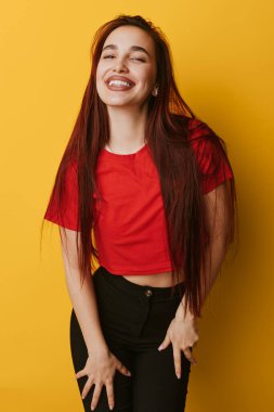 Kırmızı bluzlu, uzun düz saçlı, gülümseyen genç bir kadın ve elleri dizlerinin üzerinde poz veren siyah bir pantolon. Parlak sarı arkaplan ve sıradan moda çekimlerinde neşeli bir ifade..