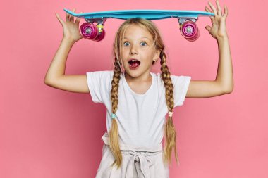 Uzun örgülü, gülümseyen genç bir kız başının üstünde parlak mavi bir kaykay tutuyor. Pembe arka plan ve günlük beyaz kıyafet neşeli ve neşeli bir hava yaratır..