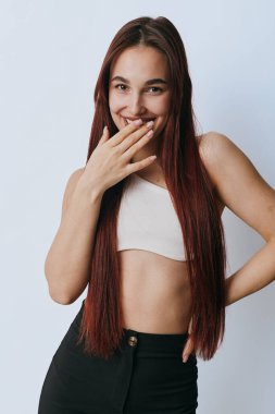 Uzun saçlı, ağzı beyaz tişörtlü ve arka planda duran siyah pantolonlu gülümseyen genç bir kadın. Doğal, neşeli, rahat ve rahat bir poz..