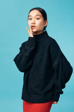 Siyah süveter ve kırmızı etekli genç bir kadının portresi. Mavi arka planda eli yüzüne yakın poz veriyor. Stüdyo ortamında sakin ve yansıtıcı ruh haliyle kameraya bakıyor..