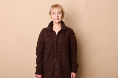 Sade bej renkli bir kazak giyen kısa sarı saçlı sakin, son sınıf öğrencisi bir kadın bej bir duvara dayanıyor. Yaşlanma, güven, moda ve günlük tarz portresi kavramı.