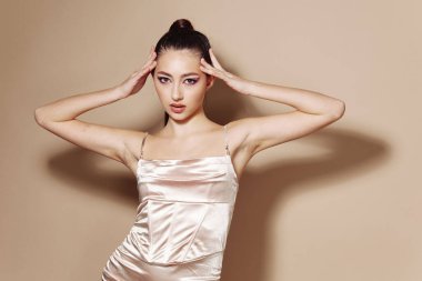 Makarna askılı parlak saten bir elbise giyen şık genç bir kadın dramatik bir poz vererek kafasına dokunuyor, bej renkli bir stüdyo arkaplanında kendine güvenen makyajla kameraya bakıyor..