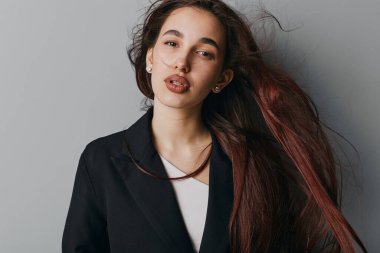 Siyah ceketli ve beyaz gömlekli, dalgalı saçlı ve minimum makyajlı bir kadın model. Stüdyoda duruyor. Tarz ve zarafet için gri arka planı var..
