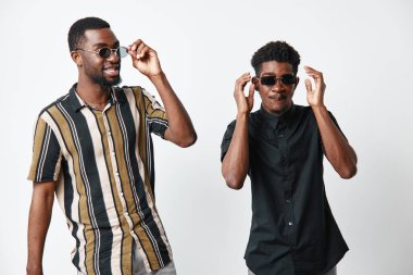 Siyah tenli, günlük giysiler ve güneş gözlüğü takmış, beyaz arka planda durup gülümseyen, arkadaşlık ve yaşam tarzı portresi olan genç siyahi adamlar..