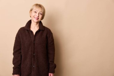 Samimi kahverengi ceket giyen mutlu olgun bir kadın bej duvarın önünde poz veriyor, içten bir şekilde gülümsüyor. Gündelik moda, orta yaşlı yaşam tarzı, rahat kıyafetler ve pozitif ruh hali kavramı.