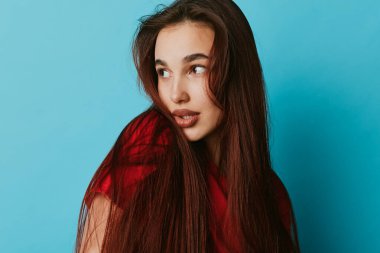 Uzun düz saçlı ve kırmızı bluzlu genç bir kadının portresi mavi arka plana bakıyor. Modern fotoğrafçılıkta rahat bir ifade ve doğal bir görünüme sahip bir kadın..