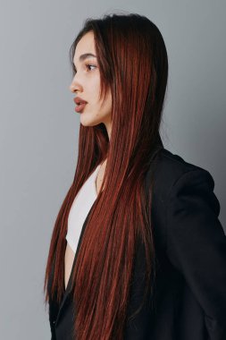 Uzun kahverengi saçlı, siyah ceketli ve beyaz gömlekli, gri arka planda duran genç bir kadın profili. Profesyonel fotoğraf çekiminde sakin yüzlü ve doğal görünüşlü bir kadın model..