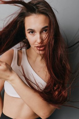 Dalgalı kahverengi saçlı ve beyaz kolsuz bluzlu genç bir kadının portresi, kendine güvenen bakışlar ve doğal bir bakışla stüdyoda nötr gri arka plana karşı poz veriyor..