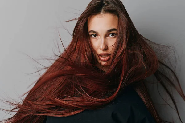 Havada uçuşan canlı kızıl saçlı genç bir kadın ve gri arka planda poz veren yoğun bakışlar. Stüdyoda saç dokusu ve yüz özelliklerine odaklanmış yakın plan portre.