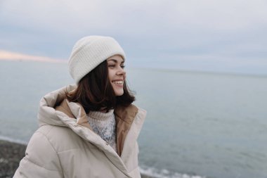 Bej ceketli ve beyaz bereli gülümseyen kadın denizin kenarında duruyor, serin havanın ve kıyı havasının tadını çıkarıyor. Sakin kompozisyon açık hava modasını ve soğuğa karşı dayanıklılığı vurguluyor
