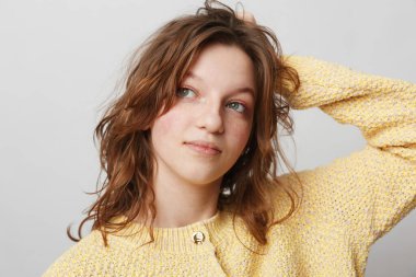 Sarı örülü süveter giyen dalgalı saçlı genç bir kadının portresi başka tarafa bakıyor. Tarafsız gri arka plan, doğal ışık, gündelik moda ve stüdyo ortamında rahat bir ruh hali..