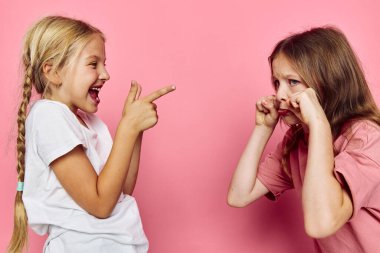 İki genç kız pembe bir arka plan önünde şakalaşıyorlardı. Bir kız gülüyor ve işaret ediyordu. Diğeri ise parmaklarıyla ağlıyormuş gibi yapıyordu..