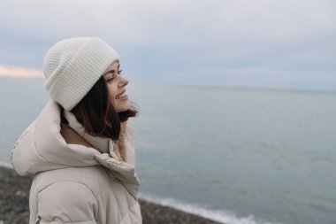 Kışlık ceketli ve örgü şapkalı bir kadın denize bakıyor. Hafif bulutlu bir ışık, açık ufuk ve huzurlu bir ruh hali içinde sakin bir kıyı portresi için siluetini geliştiriyor..